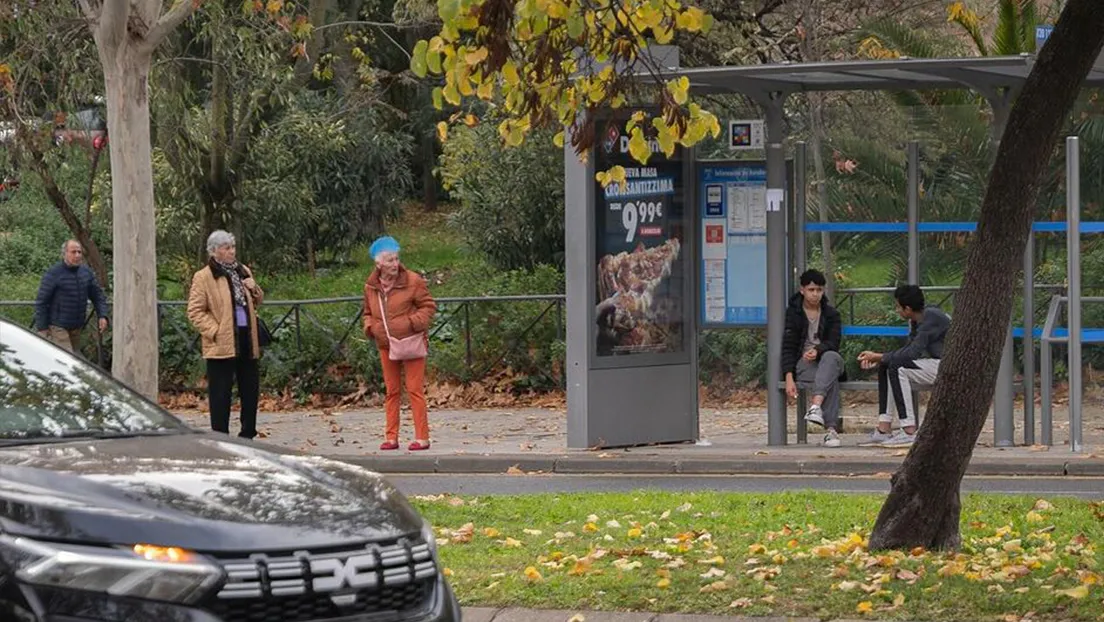 Dos mujeres mayores observan intranquilas y de pie a dos menas que están sentados en la parada del autobús